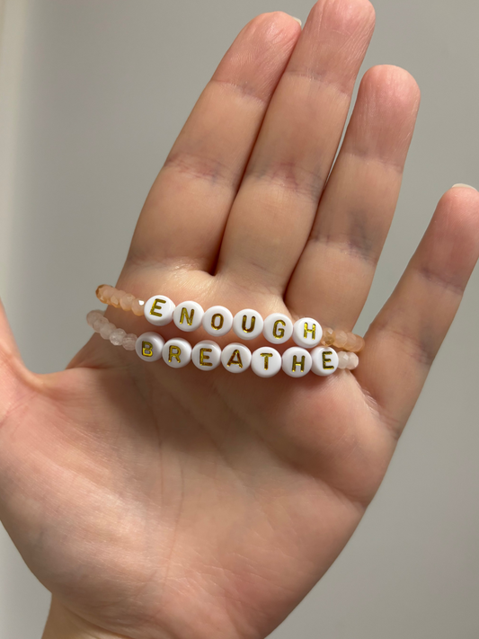 A hand holding two beaded bracelets. One bracelet features white round letter beads spelling "BREATHE" with gold lettering, and the other is a translucent peach-beaded bracelet spelling "ENOUGH"