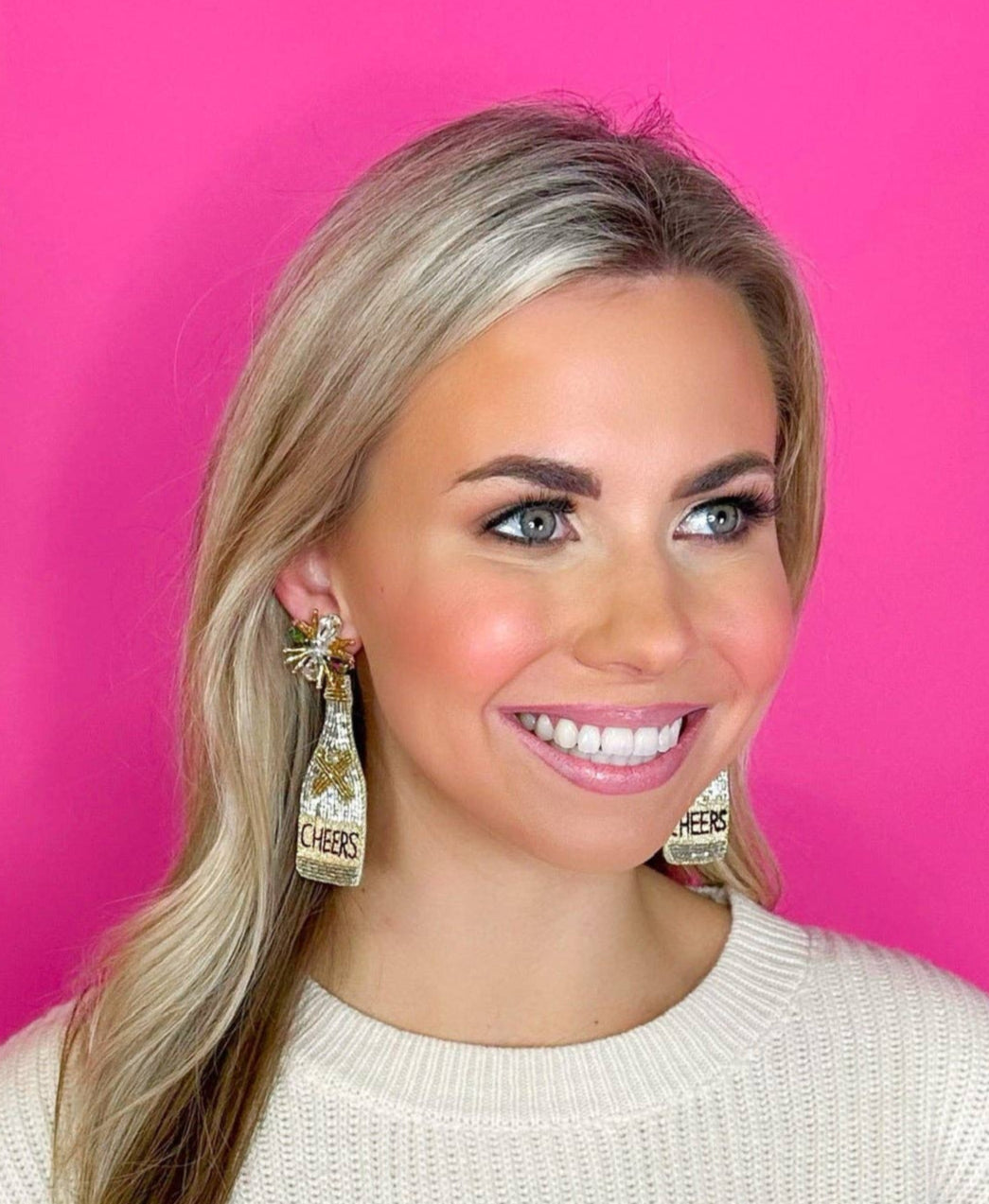 Woman wearing a white sweater with colorful sequin details against a pink background wearing large champagne bottle earrings that say "Cheers"