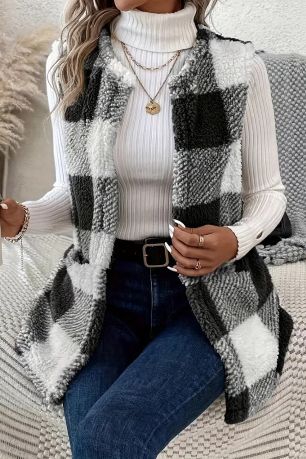 A woman wearing a white turtleneck sweater and a Sherpa plaid open front vest coat in a checkered pattern. She is also accessorized with a necklace and has a ring on her finger.