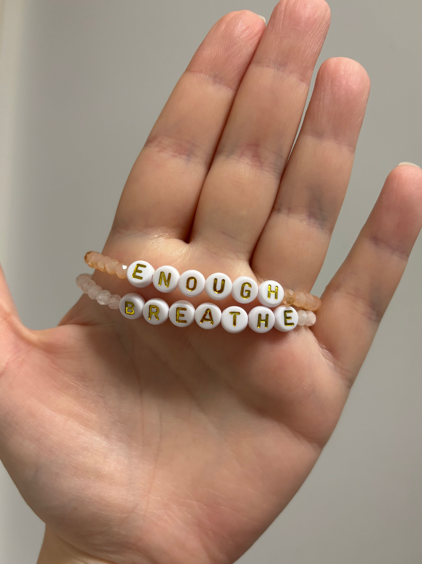 A hand holding two beaded bracelets. One bracelet features white round letter beads spelling "BREATHE" with gold lettering, and the other is a translucent peach-beaded bracelet spelling "ENOUGH"