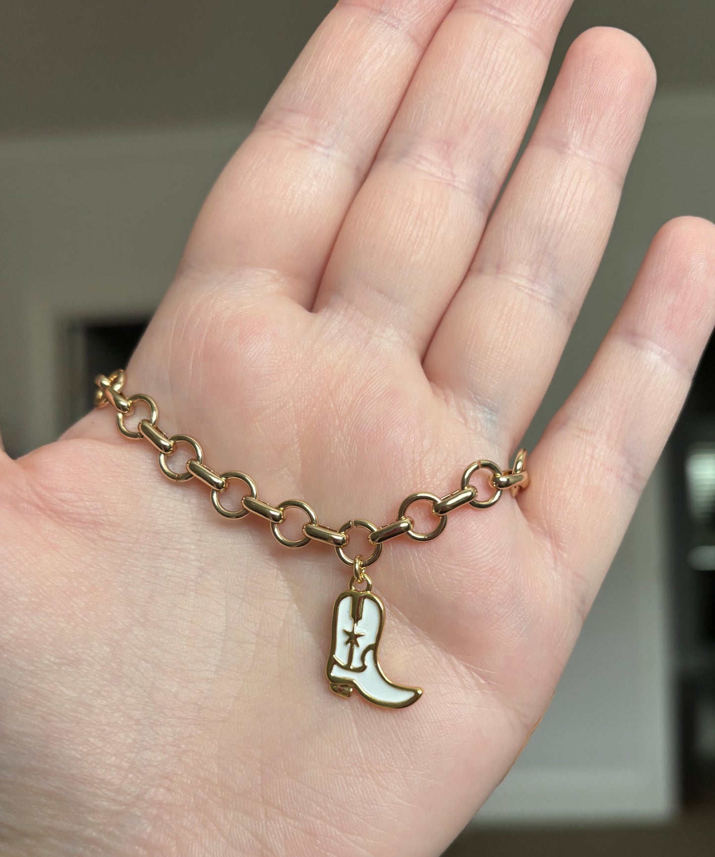 A white cowgirl boot charm on a gold-colored charm bracelet displayed on a person's hand.
