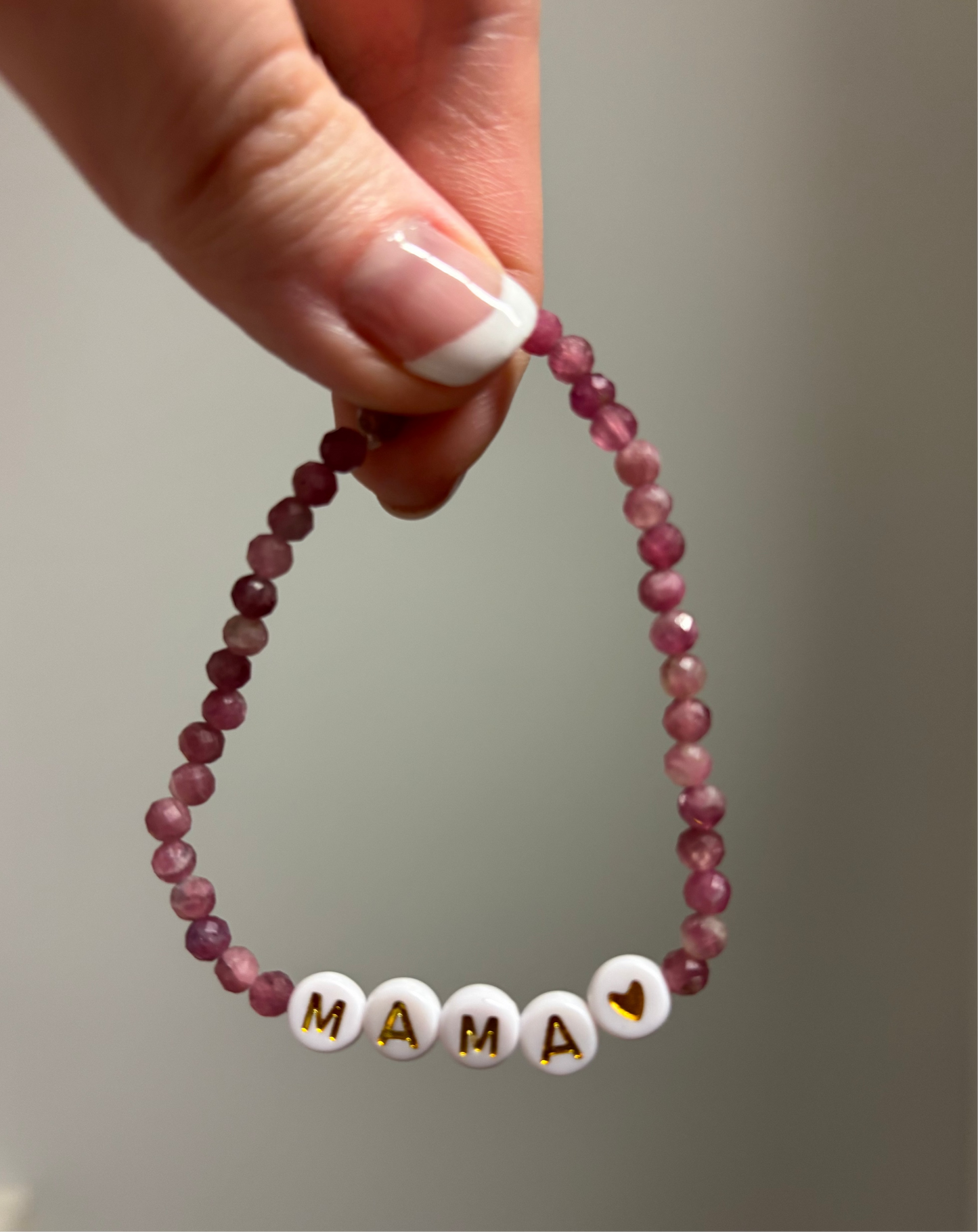 Beaded bracelet with 'MAMA' and heart charm held by a hand against a neutral background