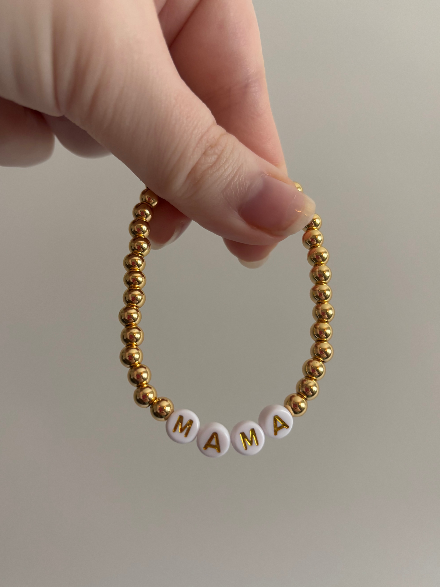 Gold beaded bracelet with 'MAMA' letters held by a hand against a neutral background