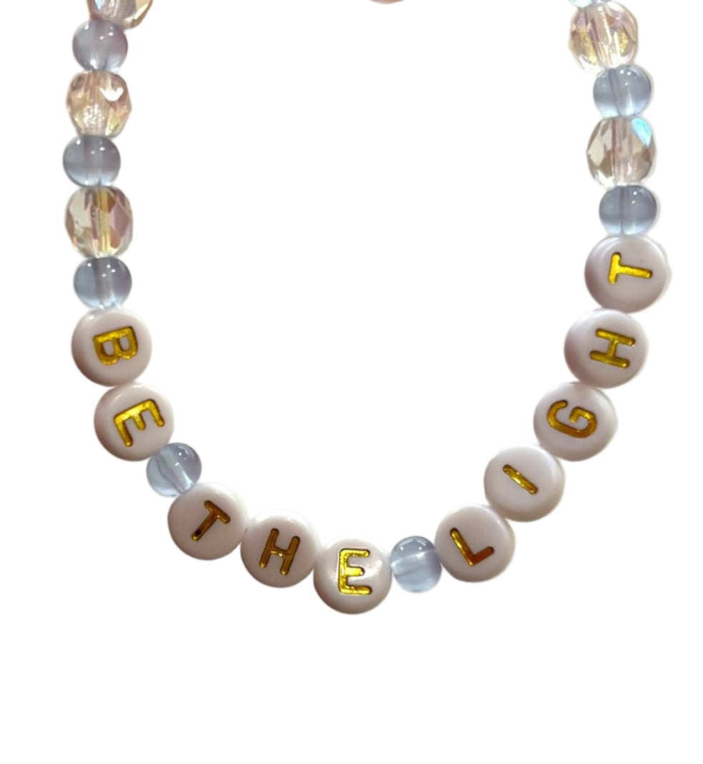 Blue & white beaded bracelet with letter beads spelling "Be the Light" held by a hand against a neutral background
