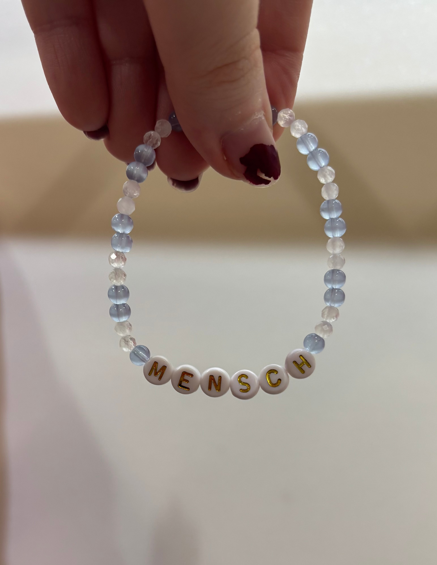 Light blue & crystal white beaded bracelet with 'MENSCH' beads held by a hand against a neutral background
