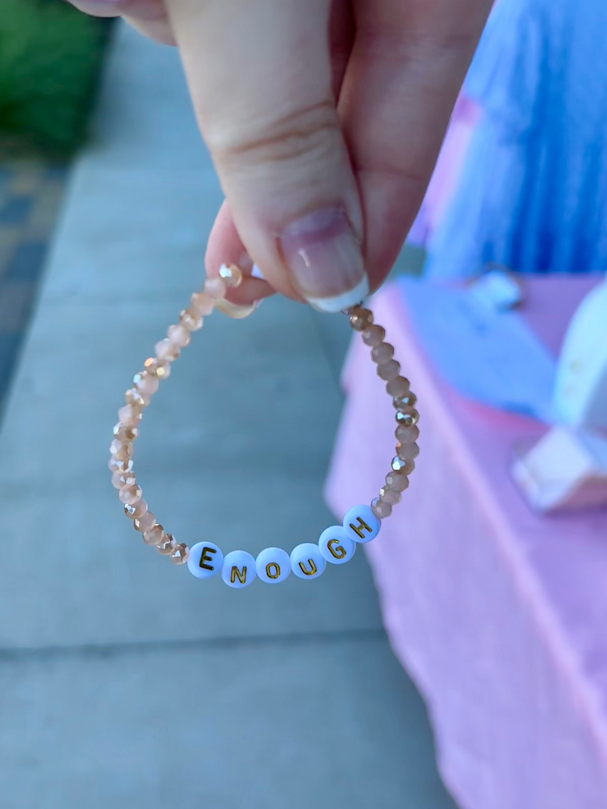 A hand holds a beaded bracelet with peach-colored faceted beads and white letter beads spelling 'ENOUGH' with small gold outlines.