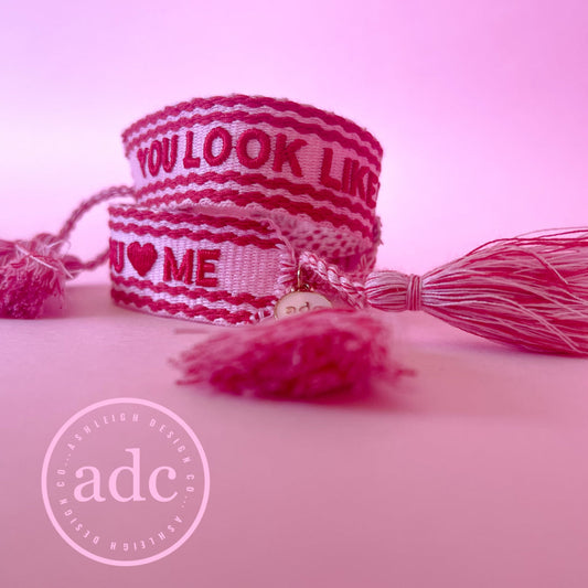 A pink woven canvas bracelet with the text 'YOU LOOK LIKE YOU LOVE ME' and a heart symbol, displayed on a pink background with a tassel detail.