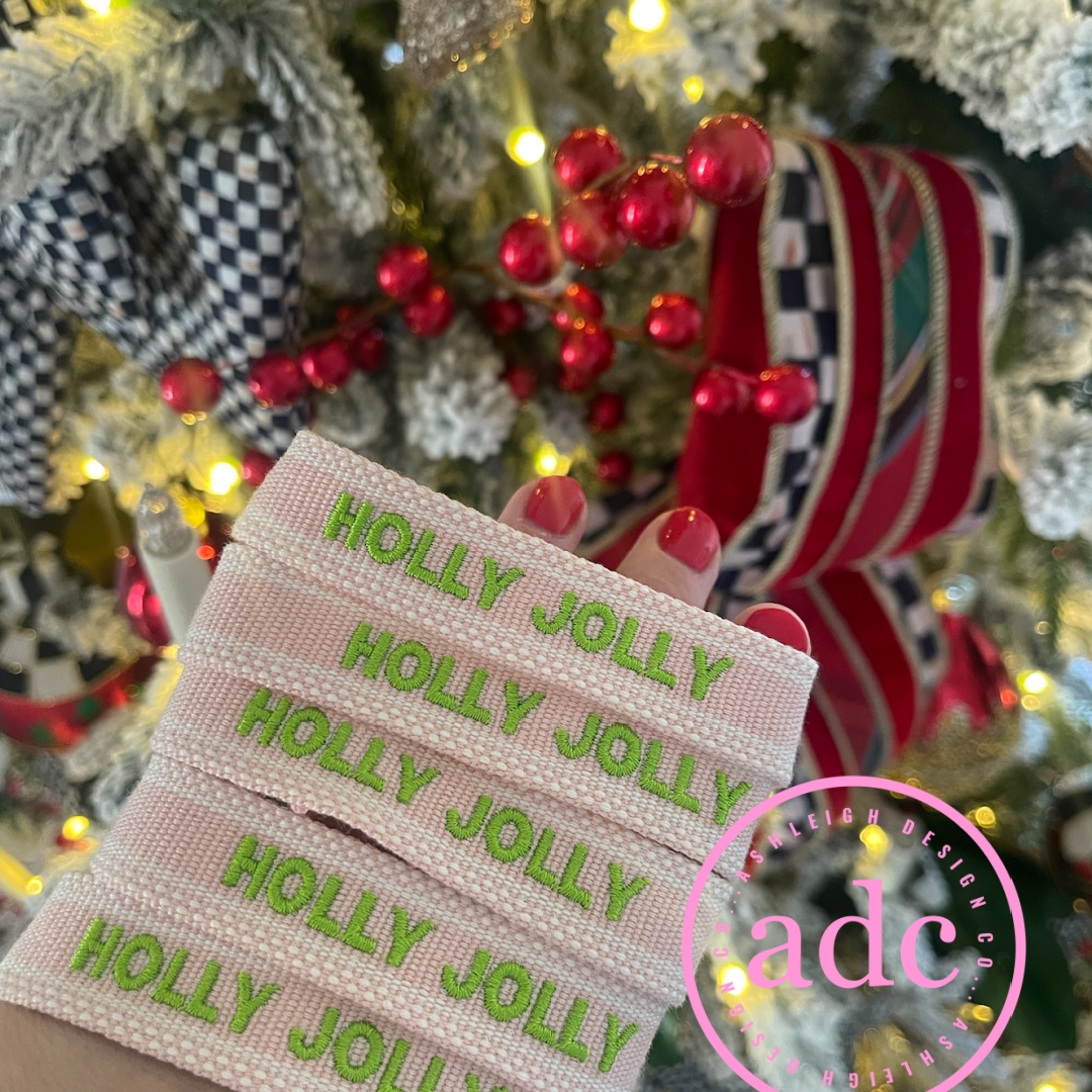 Pink adjustable woven bracelets with 'Holly Jolly' pink & green text in front of a decorated Christmas tree.