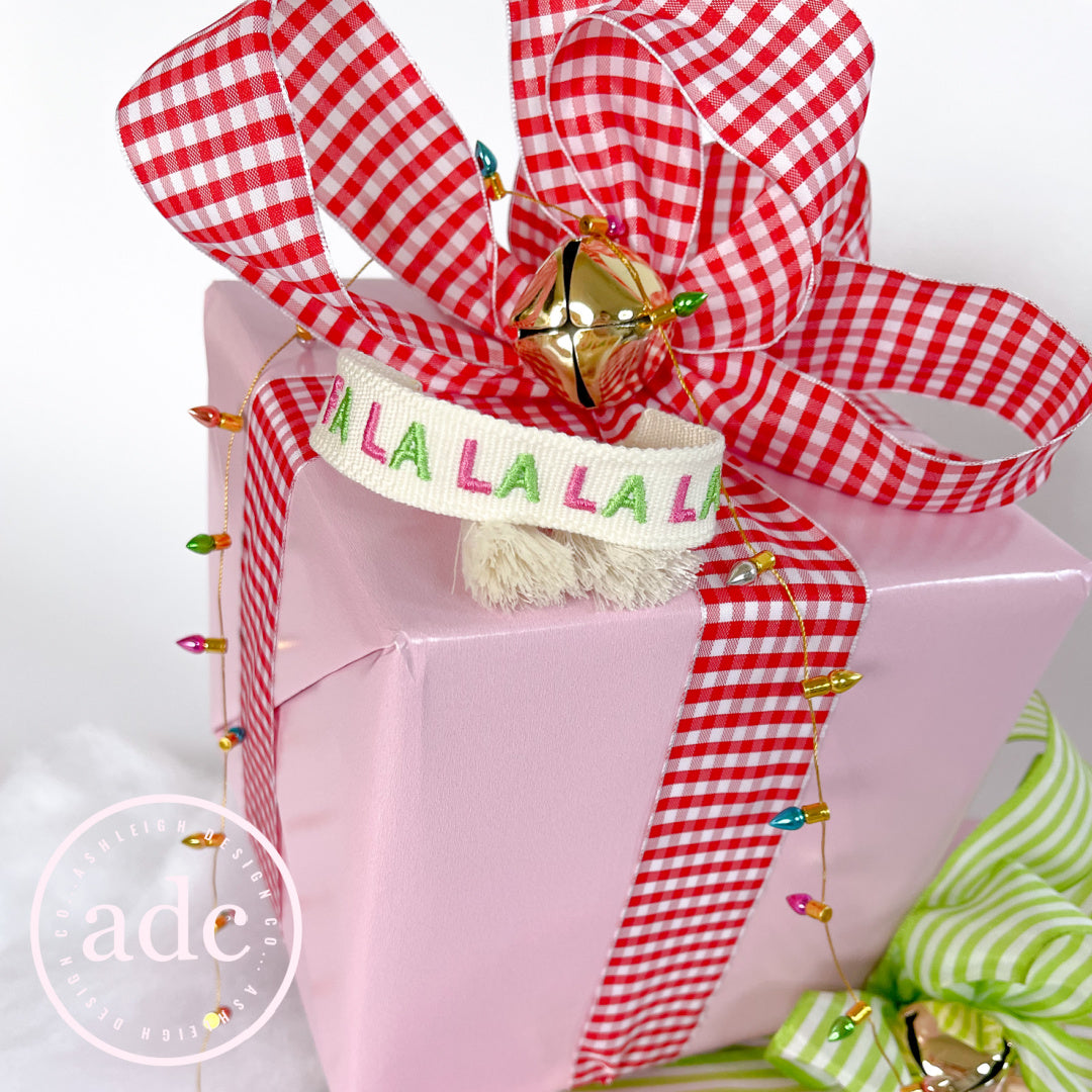A hot pink, green, and white bracelet made of woven material with 'FA LA LA' text on a present.