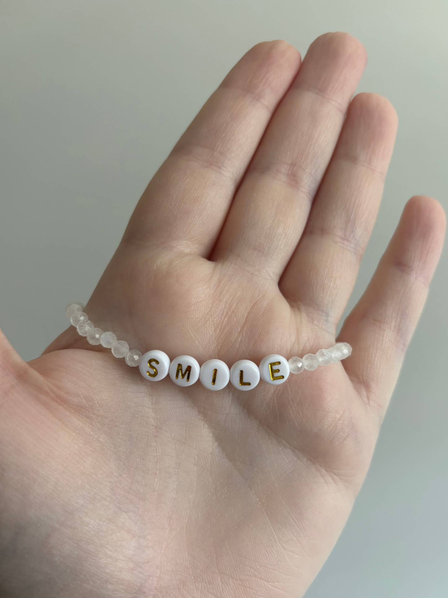 Hand holding 4mm beaded bracelet spelling "SMILE" in white beads with gold lettering