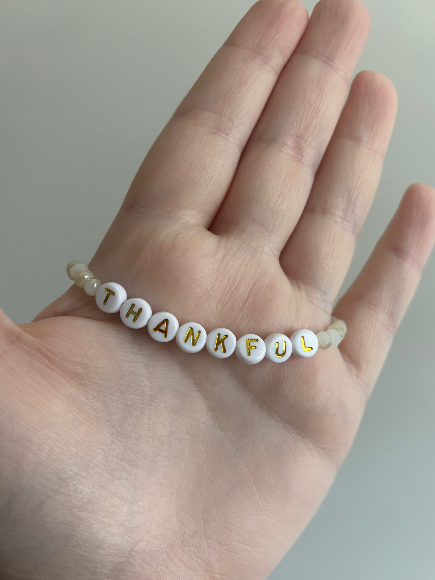 A beaded bracelet spelling 'THANKFUL' in gold lettering on white round beads, worn on the back of a person's hand.
